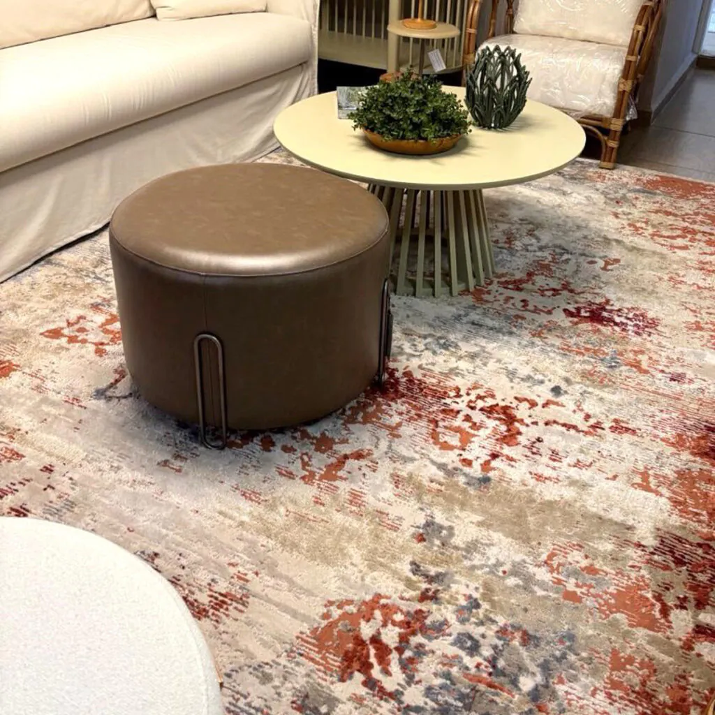 Tapete com estampa artística em tons ferrugem e neutros, instalado em sala com pufe redondo e mesa de centro clara, solução personalizada da Copato